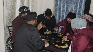 A group of people gathered around a table with food, in a room with curtains and a metal chair. - VEDA INN (Kalsi)