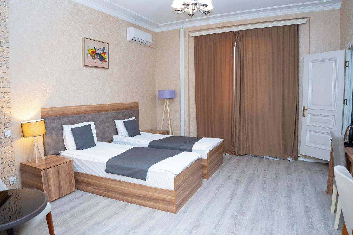Une chambre d’hôtel avec deux lits, un bureau, une chaise, une lampe, un tableau et une grande fenêtre avec des rideaux.