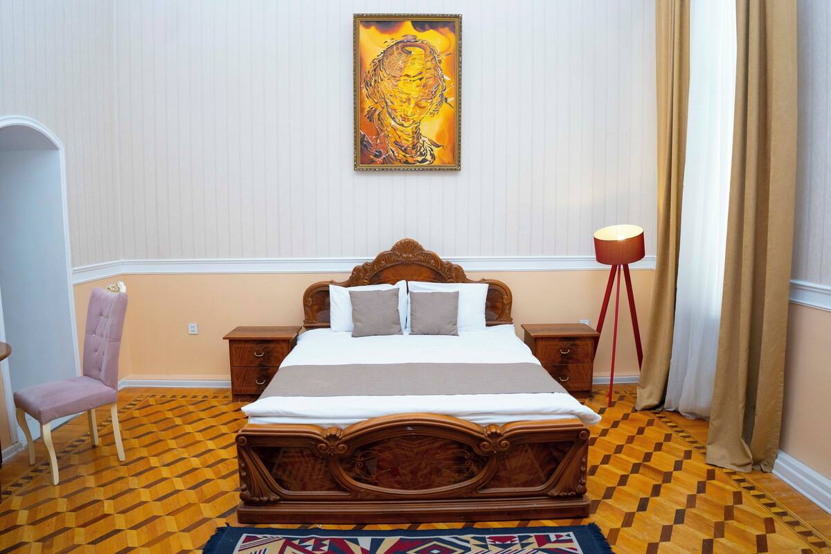 Un lit bien fait, avec une tête de lit en bois, une table de chevet, une chaise et une lampe sur pied, dans une pièce dont le sol est recouvert d’un motif décoratif et où un tableau est accroché au mur.