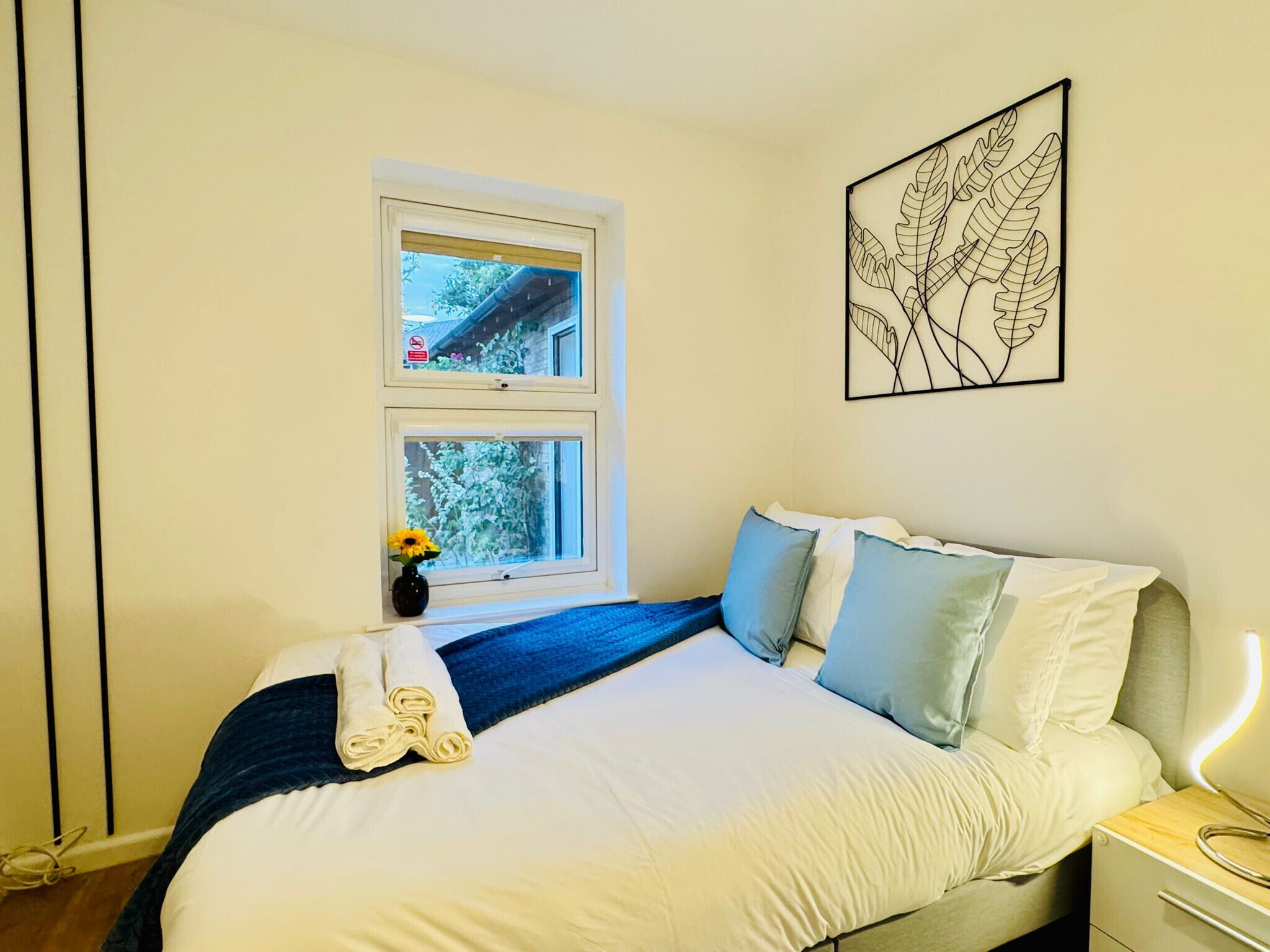 Un dormitorio con una cama, una mesita de noche, una lámpara de pared, una obra de arte enmarcada y dos ventanas.