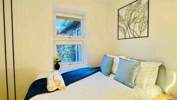 Un dormitorio con una cama, una mesita de noche, una lámpara de pared, una obra de arte enmarcada y dos ventanas.