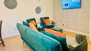 Ein modernes Wohnzimmer mit türkisfarbenen Sofas, orangefarbenen Kissen, einer runden Wanduhr, einem Flachbildfernseher und einer Wandtapete mit Muster.