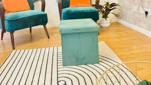 Ein Zimmer mit zwei hockerartigen Sesseln in Türkis, einem Türkis-farbenen Hocker, einem gemusterten Teppich, einer Topfpflanze und einer strukturierten Wand.