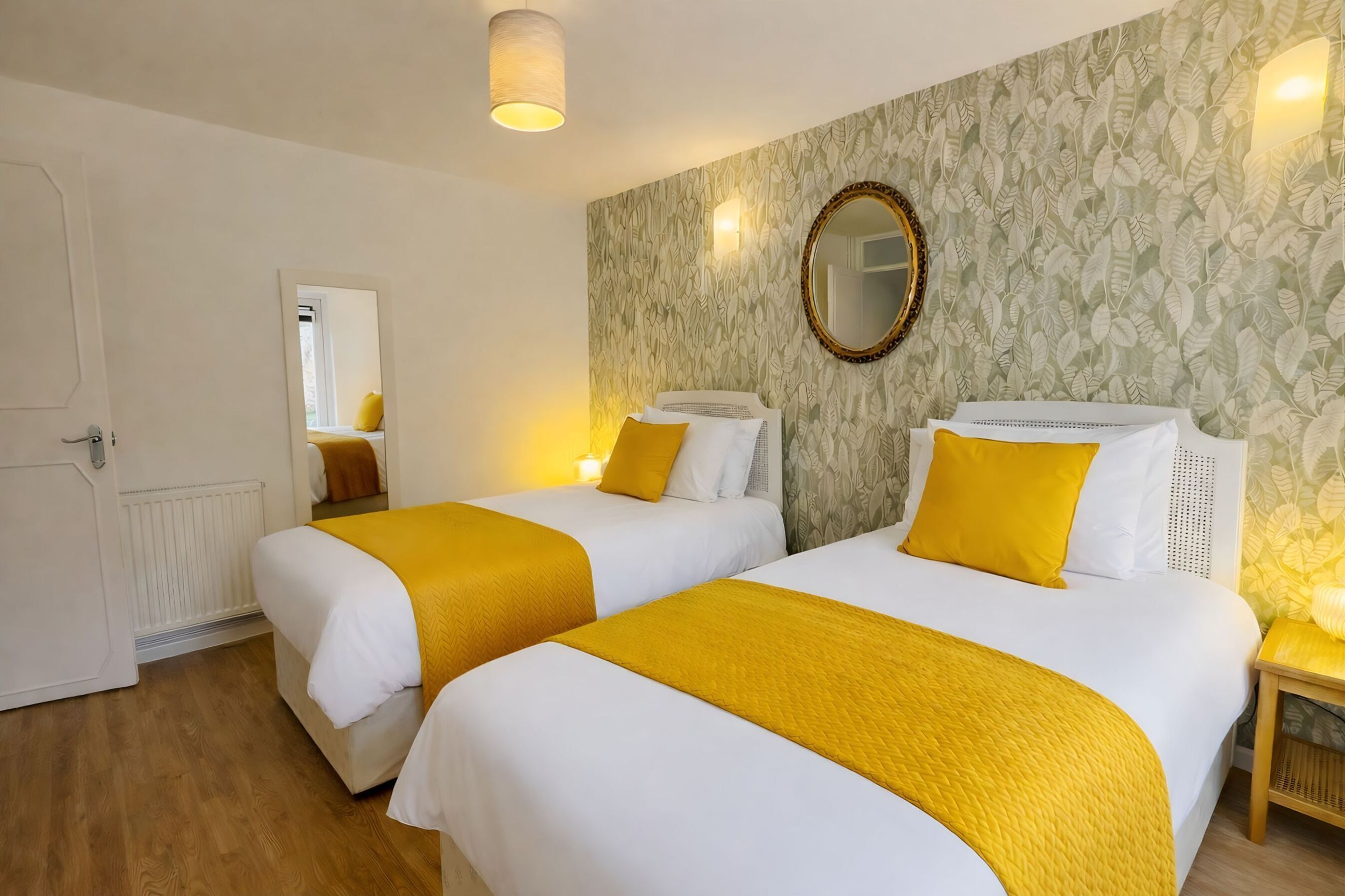 Une chambre d’hôtel avec deux lits, chacun agrémenté de linge de lit blanc et de touches de couleur jaune, un miroir au mur et un papier peint à motifs.