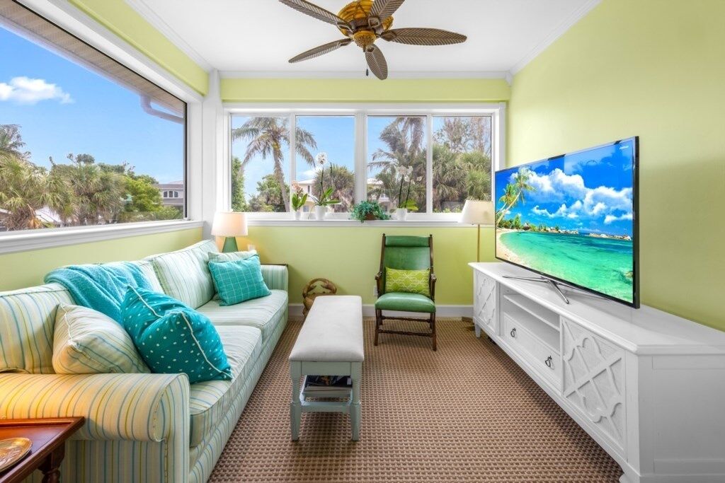 A room with a large window, a ceiling fan, a television, a green chair, a striped sofa, and a small table.