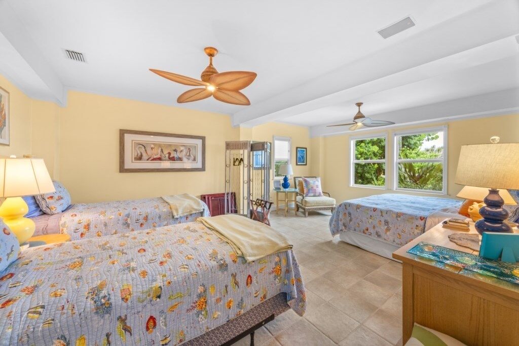 A bedroom with two beds, a ceiling fan, a desk with a lamp, and a window with a view of greenery.