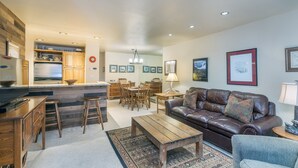 A spacious living area with a brown leather sofa, wooden coffee table, and a dining table with chairs. A kitchen with wooden cabinets and a refrigerator is visible in the background. - Alpine Escape 1 Bedroom Condo (Telluride)