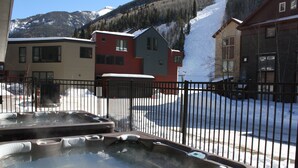 A hot tub area with a metal fence, a building with a red and gray facade, and a snowy mountain backdrop. - Alpine Escape 1 Bedroom Condo (Telluride)