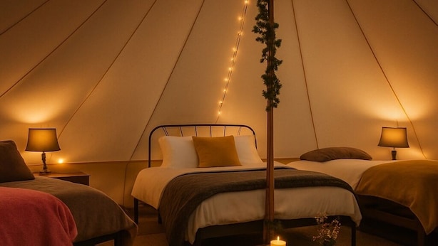 A cozy tented accommodation with two beds, each with a bedside table and lamp, a central table with a candle, and a fur rug on the floor.