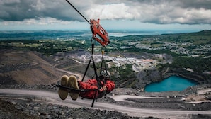 A person suspended in a harness, attached to a cable, with a mountainous landscape and a body of water in the background.