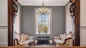 A formal room with two ornate chairs, a central table, and a large window with a view of trees.
