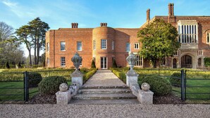 A large, historic brick building with a symmetrical facade, a central entrance with steps, and a formal garden with a gravel path leading to the entrance.