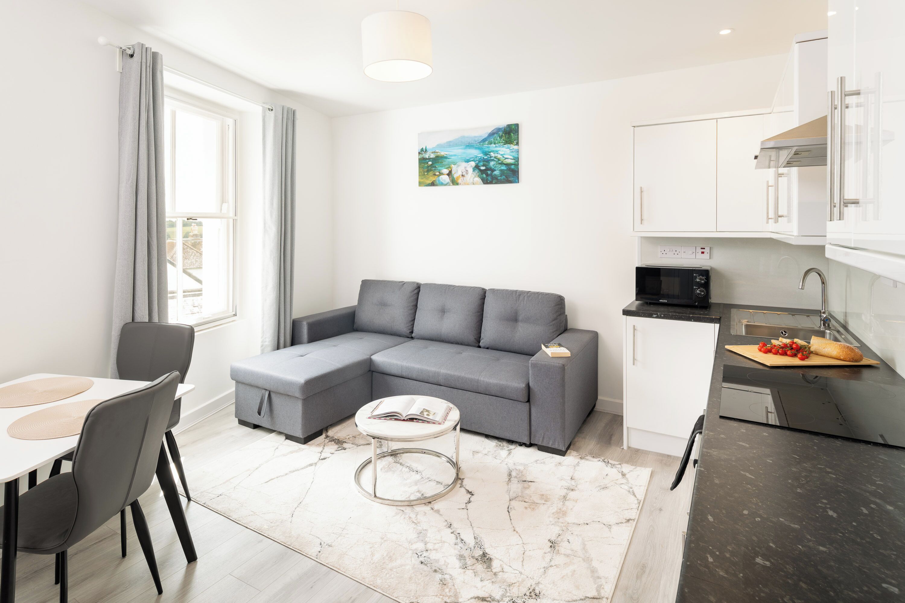 Een moderne woonkamer met een grijze modulaire bank, een ronde salontafel en een eethoek met een witte tafel en grijze stoelen. Achtergrond: een keuken met witte kastjes en een zwart aanrecht.