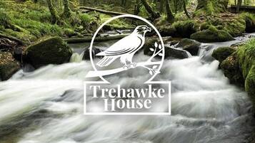 Ein Logo mit einem Vogel auf einem Ast und dem Schriftzug "Trehawke House" über einem WaldBach.