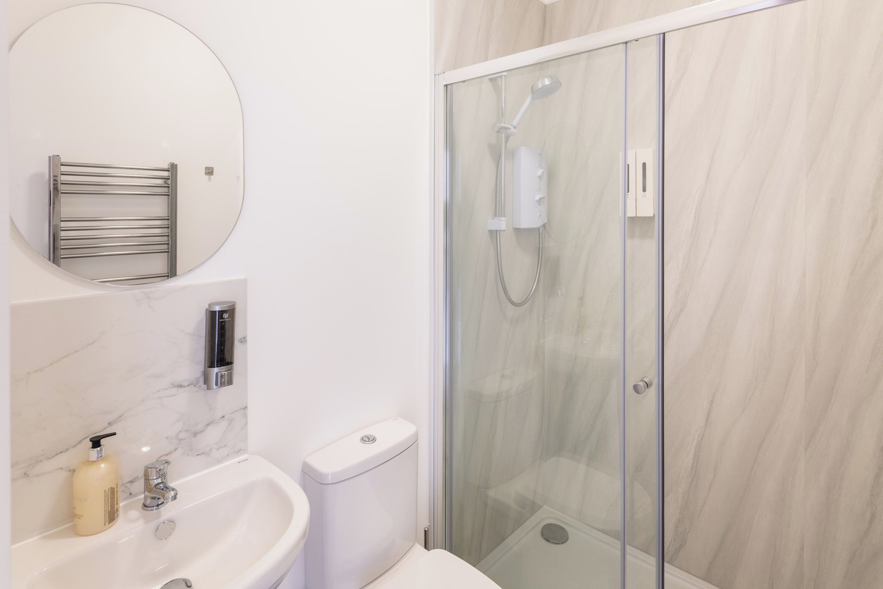 A modern bathroom with a glass-enclosed shower, a round mirror, a toilet, and a sink with a marble backsplash.