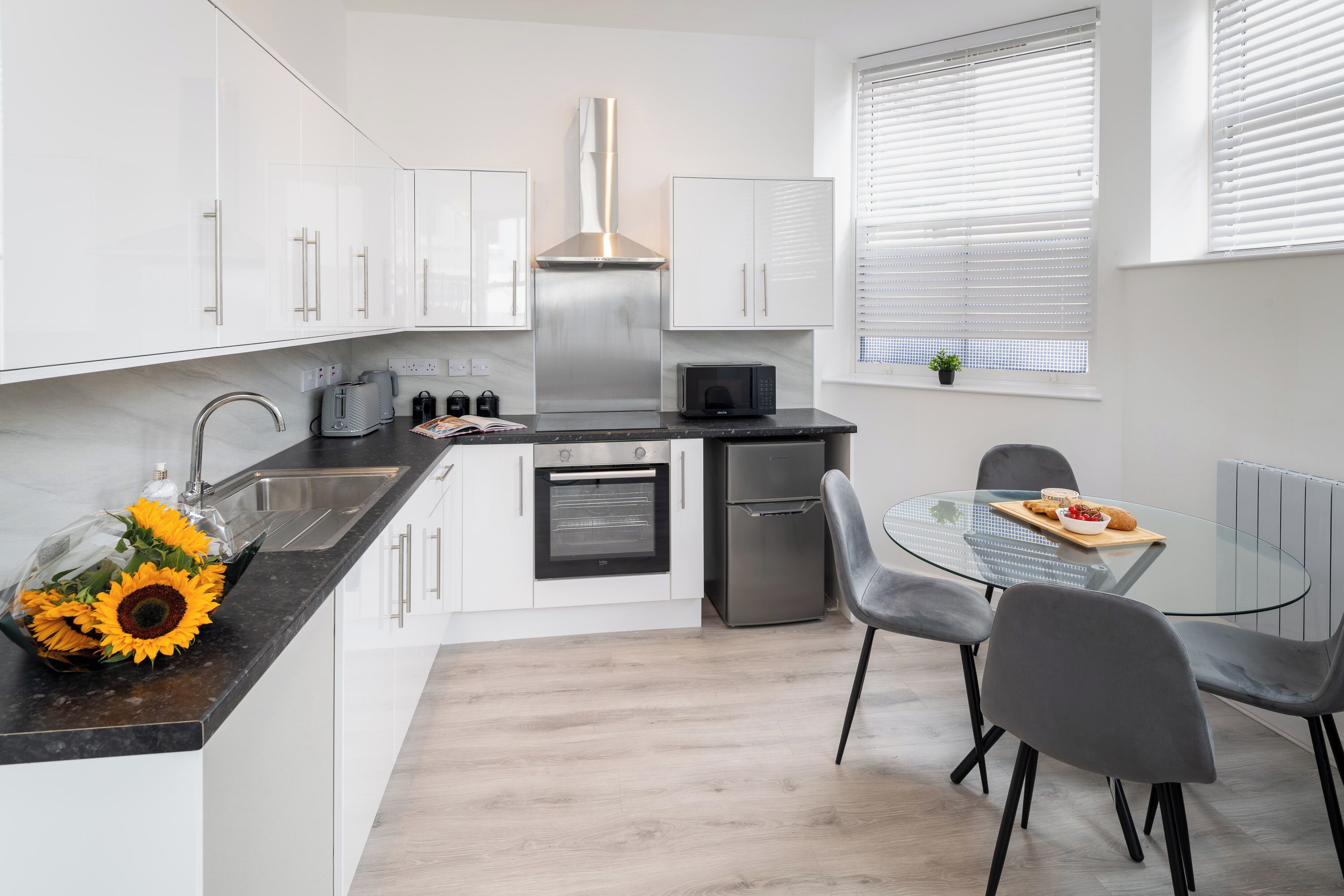 Une cuisine moderne avec des meubles blancs, des appareils en acier inoxydable et une table à manger en verre, assortie de chaises grises.