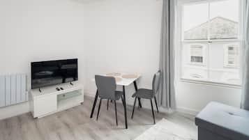Una sala de estar moderna con un televisor de pantalla plana, una mesa de comedor con sillas, un sofá gris y una ventana con cortinas.