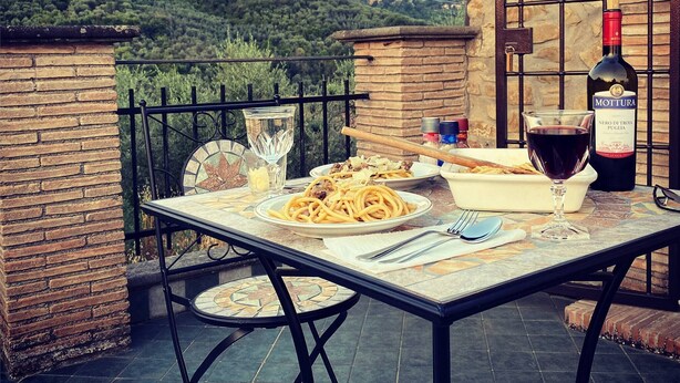 Une table à manger en plein air, avec des assiettes remplies de nourriture, une bouteille de vin et des verres, sur une terrasse offrant une vue sur une colline.