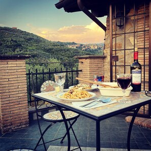 Ein Essplatz im Freien mit Speisen, einer Weinflasche und Gläsern auf einer Terrasse mit Blick auf eine Hügelkuppe.