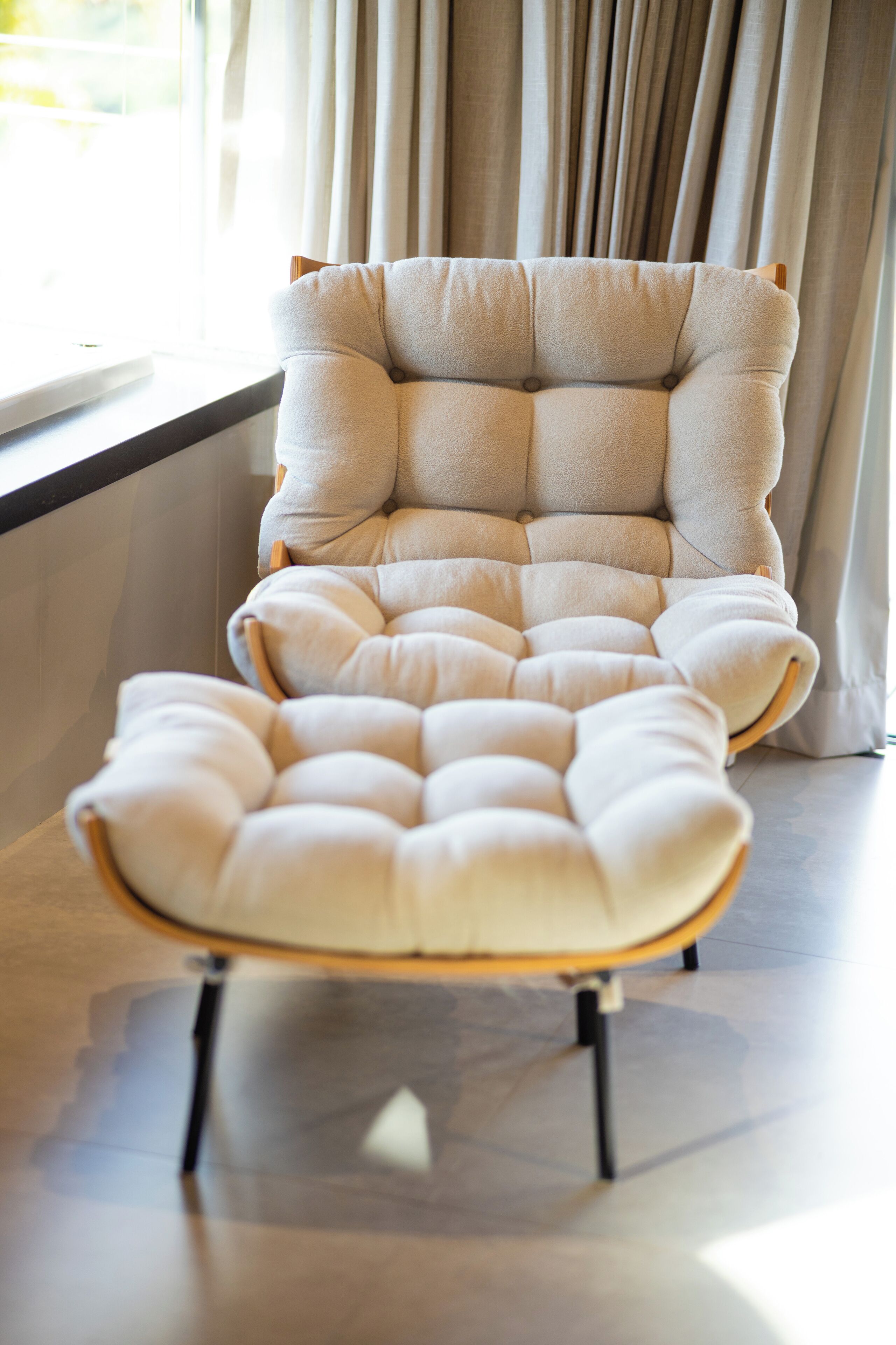 A tufted armchair with wooden legs in a room with a large window and curtains.
