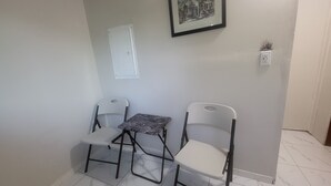 Une petite pièce avec un sol carrelé, deux chaises blanches, une petite table, un tableau encadré accroché au mur et une porte.