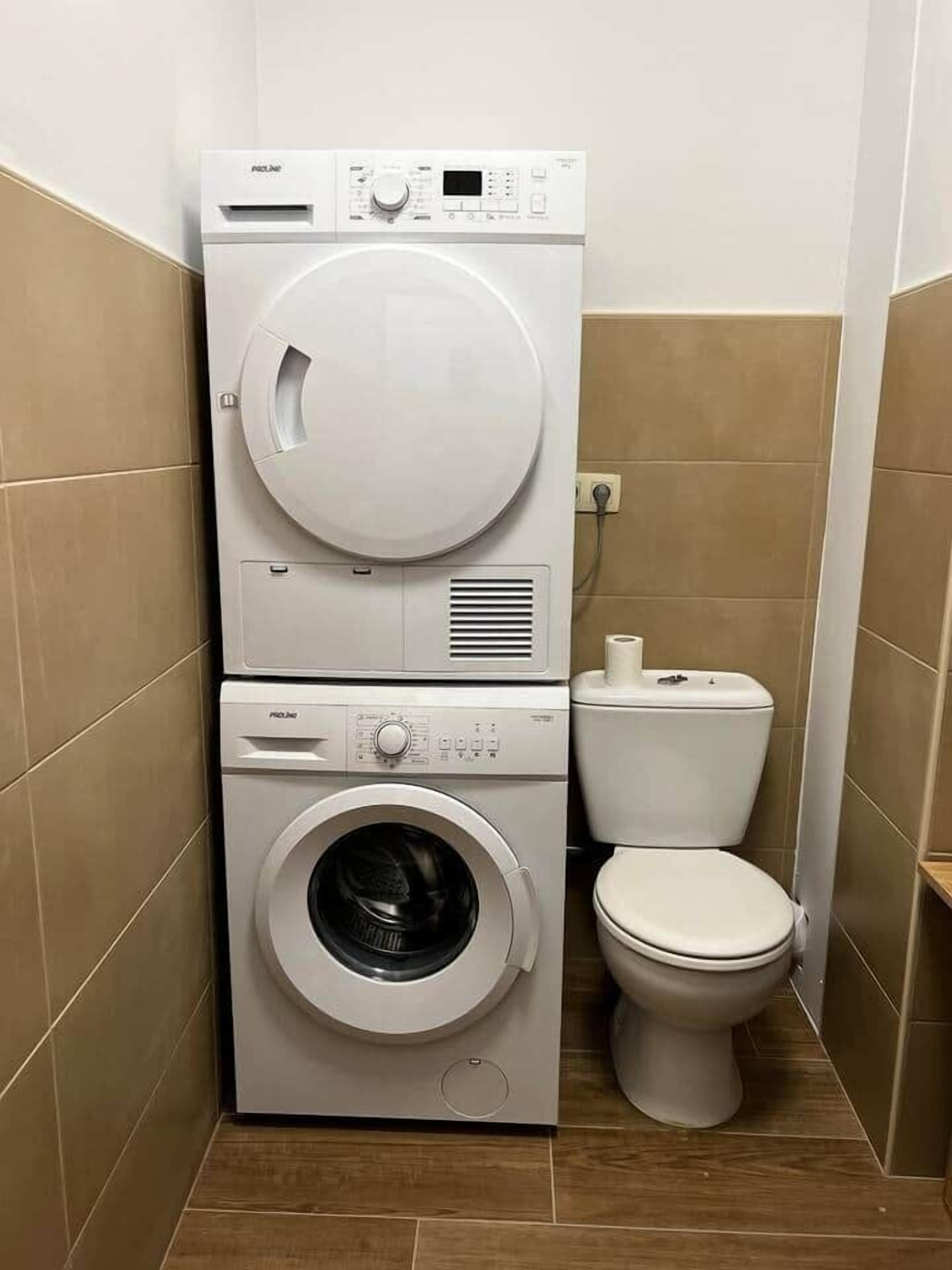 Une petite buanderie équipée d’un lave-linge et d’un sèche-linge superposés, de toilettes et de murs carrelés.