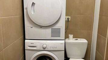 Une petite buanderie équipée d’un lave-linge et d’un sèche-linge superposés, de toilettes et de murs carrelés.