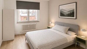 Ein Schlafzimmer mit Bett, Nachttischen, Heizkörper, Fenster mit Blick auf Gebäude und einem gerahmten Bild an der Wand.