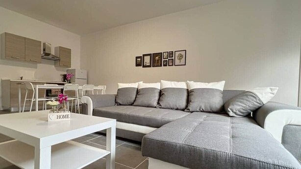 Ein modernes Wohnzimmer mit einem grauen Ecksofa, einem weißen Couchtisch und einer Küche im Hintergrund.