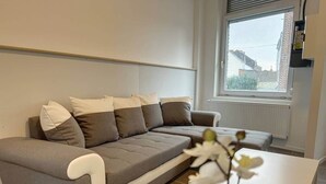 A modern living room with a grey sofa, white cushions, a white coffee table, and a window with a view of buildings.