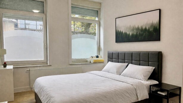 Ein Schlafzimmer mit einem Bett, einem Nachttisch, einem Fenster und einem gerahmten Bild an der Wand.