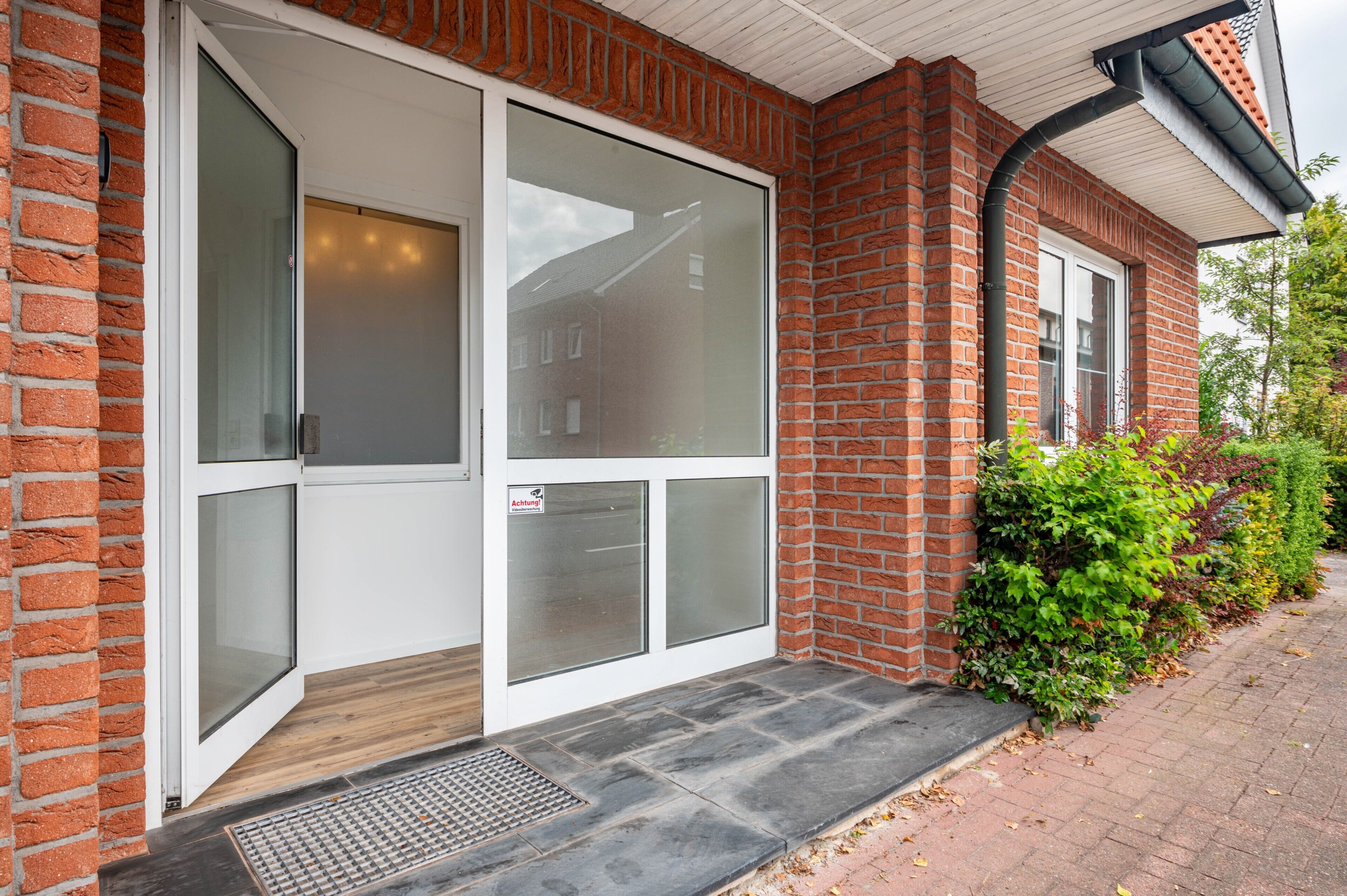 A brick building with a glass door entrance, a metal grate at the bottom, and a paved walkway.