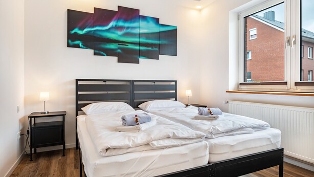 A double bed with white linens, a black headboard, two bedside tables with lamps, and a large window with a view of buildings. - Apartment 'Die Alter Schwede 1' with Private Terrace and Wi-Fi (Vechta)