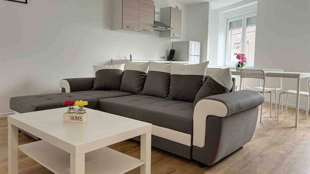 A modern living room with a grey sofa, a white coffee table, and a kitchen area in the background.