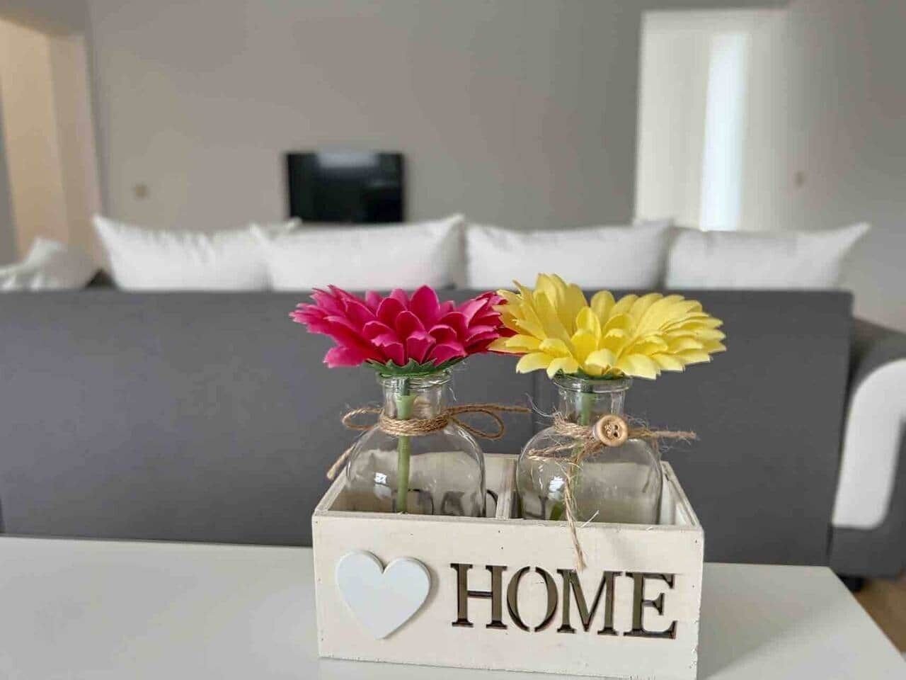 Ein weißer Tisch mit einer Holzkiste, auf der 'HOME' steht, und zwei Glasflaschen mit Blumen.