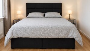A bedroom with a black headboard, two bedside tables with lamps, a bed with a patterned bedspread, and a window with curtains.