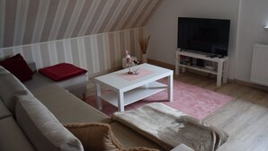 Un salon avec un plafond en pente, un téléviseur à écran plat, une table basse blanche et un tapis rose.