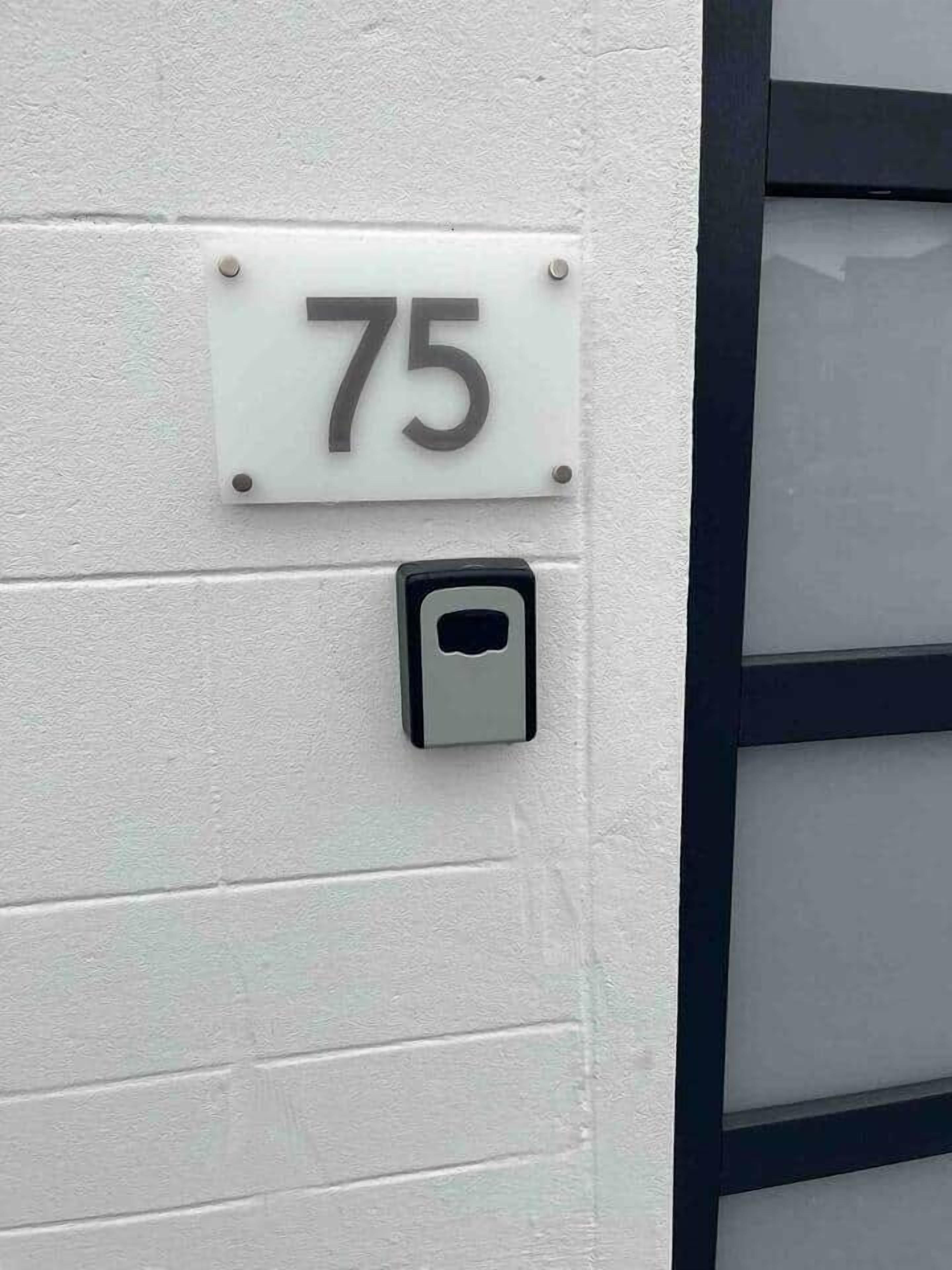 Número de casa '75' en una pared blanca, con una puerta y un teclado electrónico.