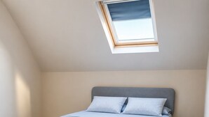 Une chambre avec un plafond en pente, un puits de lumière, un lit avec une literie grise et un plancher en bois.