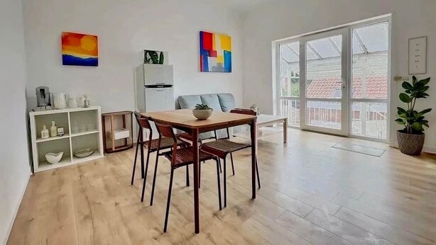 Ein modernes Wohnzimmer mit Holzboden, einem Esstisch mit Stühlen, einer Couch, einer weißen Schrankwand und einem großen Fenster mit Blick.