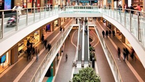 A multi-level shopping mall with a central escalator, glass railings, and various retail stores.