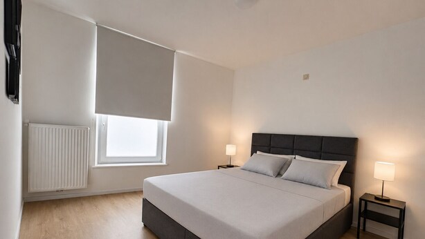 Une chambre à coucher avec un grand lit, deux tables de chevet, une fenêtre avec des stores, un radiateur et un luminaire au plafond.