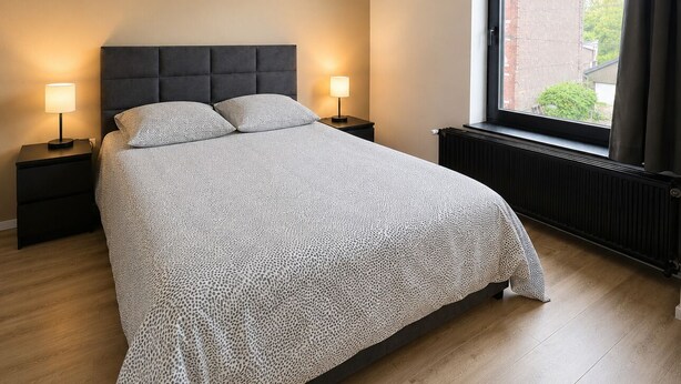 Une chambre à coucher avec un grand lit, deux lampes de chevet, une fenêtre donnant sur un immeuble et un radiateur.