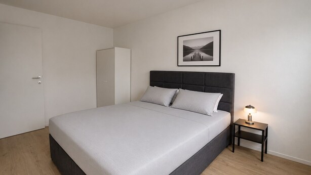 A bedroom with a bed, a nightstand with a lamp, a wardrobe, and a framed picture on the wall.