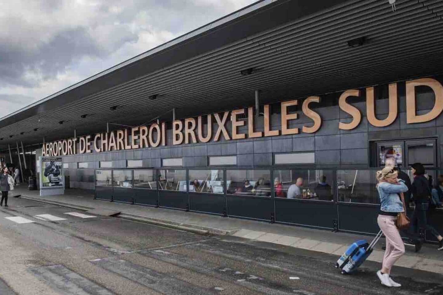Een luchthaventerminal met 'AEROPORT DE CHARLEROI BRUXELLES SUD' aanduiding, een overdekt wachtgebied en een persoon met bagage.