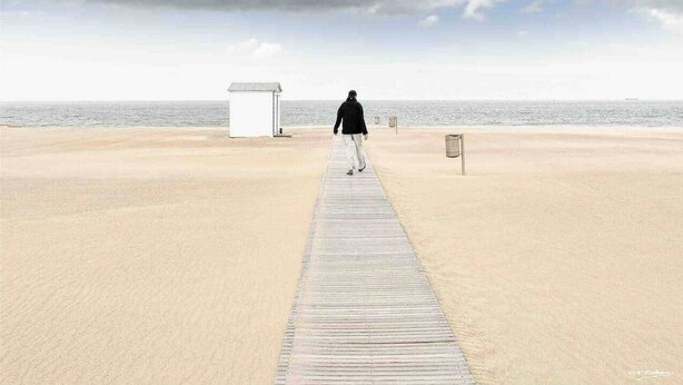 Una persona camina sobre una pasarela de madera hacia una playa bajo un cielo nublado.