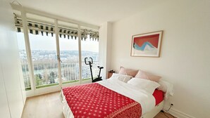 A bedroom with a large window, a bed with a red patterned bedspread, a treadmill, and a painting on the wall. - Panoramic Paris Views - 3BR Near Eiffel Tower & Seine (Boulogne-Billancourt)