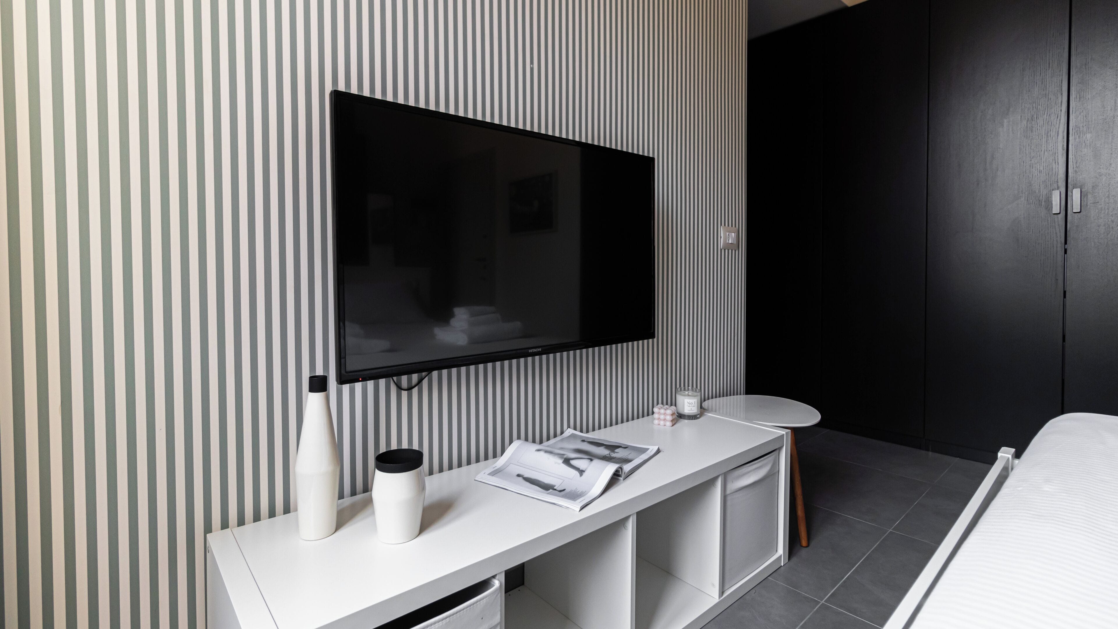 Une chambre d’hôtel moderne dotée d’un grand téléviseur à écran plat fixé au mur, d’une console blanche sur laquelle repose un magazine, de deux vases blancs et d’une petite table ronde.