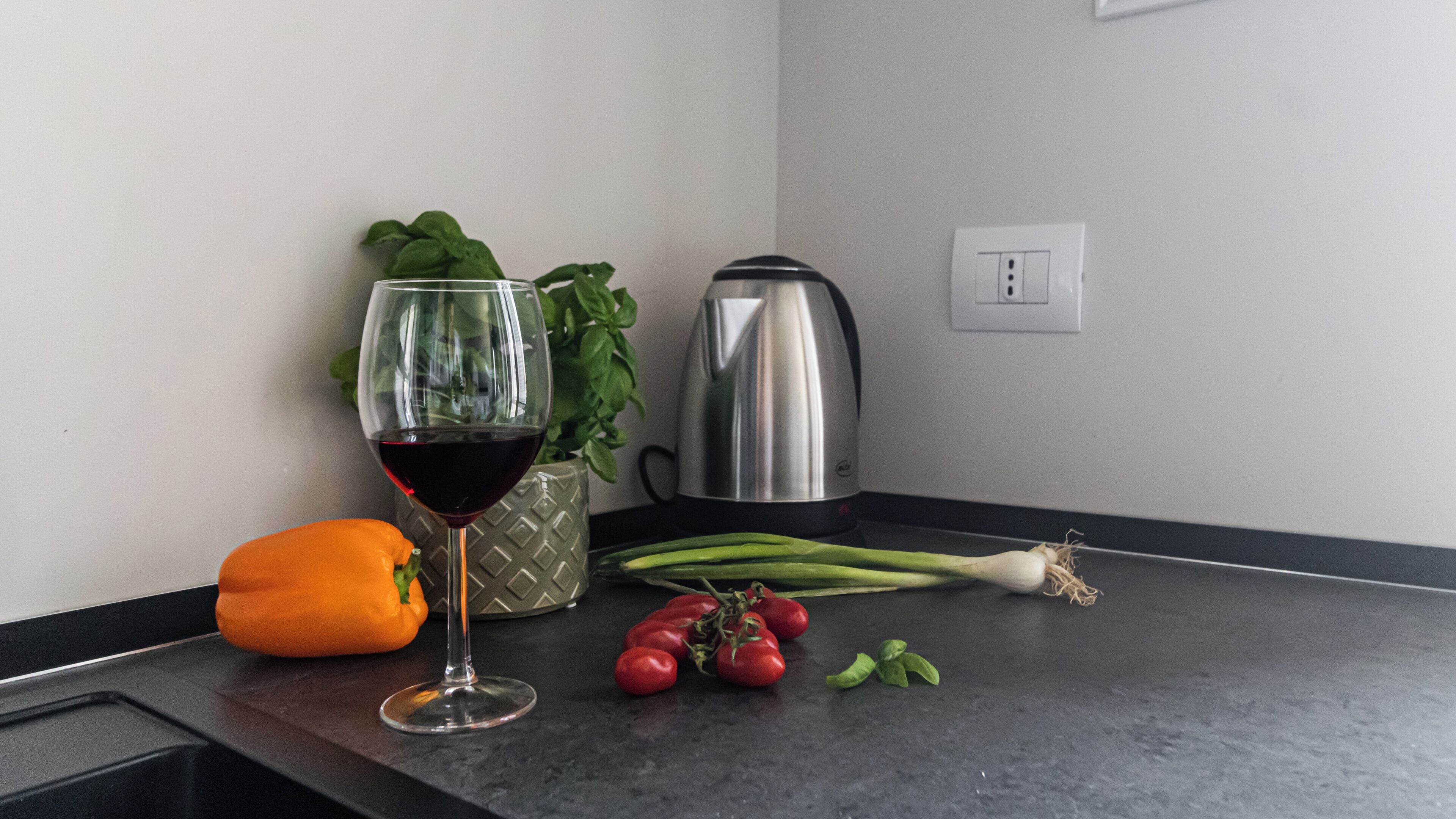 Un comptoir de cuisine avec un verre de vin rouge, un poivron, des tomates, des oignons verts et une bouilloire.