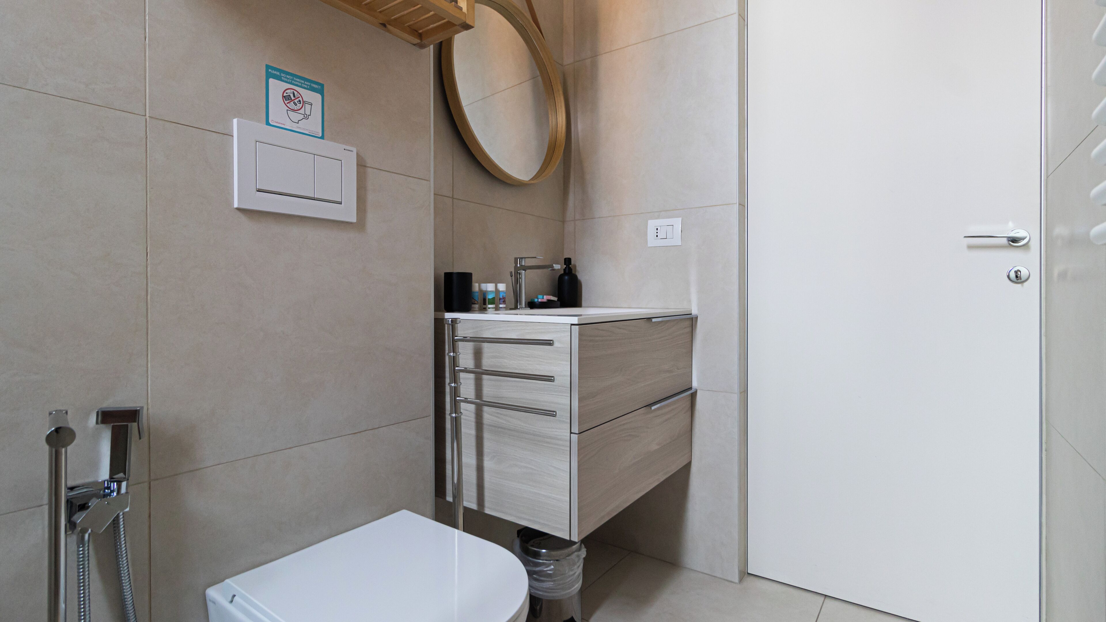 A bathroom with a toilet, sink, and shower. There is a mirror above the sink and a sign on the wall.